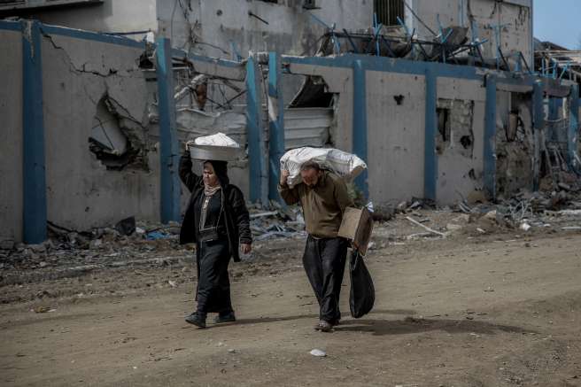 A man and woman carry supplies walking past buildings destroyed